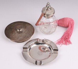 A circular silver ashtray with engine turned decoration and monogrammed, Chester 1921, 11cm, 60 grams together with a cut glass perfume atomiser with silver collar, a circular silver powder puff lid the interior set a mirror (a/f) 