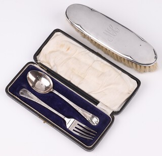 A cased silver 2 piece christening set with spoon and fork, monogrammed (slightly misshapen), Birmingham 1923, by A J Bailey,  36 grams and an oval silver clothes brush monogrammed ML, Birmingham 1917 
