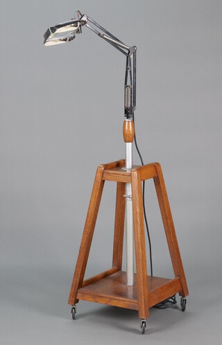 A magnifying lamp with rectangular magnifying glass flanked by 2 bulbs marked The Anglepoise PAT in UK and Abroad, raised on a tapered oak 2 tier stand with casters 145cm h x 46cm w x 45cm d 