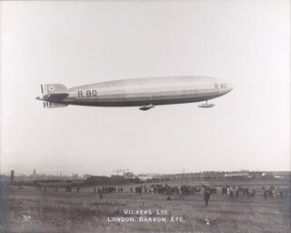 Vickers Ltd -  A side view of the R80 Airship in Flight at Walney Island, Barrow in Furness, gelatin silver print photograph, published by Vickers circa 1920, 56cm x 70cm, within a stained wood frame 66cm x 80cm. Provenance: Sir Barnes Wallis thence by descent.  Note: The R.80 was a British rigid airship, first flown on 19 July 1920, and notable as the first fully streamlined airship constructed in Britain.  Designed by Barnes Wallis and H. B. Pratt for Vickers, it was originally commissioned as a military project for the British Admiralty but was ultimately completed for commercial passenger service. In practice, however, the R.80 proved too small to operate viably in this capacity.  After a brief period assisting in the training of personnel from the United States Navy who were to crew the ZR-2 airship, the R.80 was withdrawn from service and was eventually scrapped in 1925